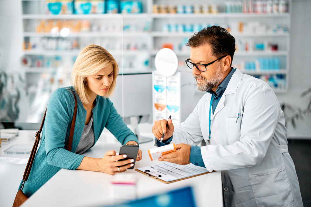 Mid adult woman using cell phone while consulting with her pharmacist in choosing medicine in a pharmacy.